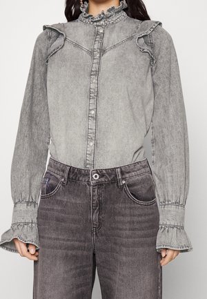 Femme portant un chemisier en denim gris délavé à l'acide avec un col montant à volants et des poignets, associé à un jean taille haute gris foncé délavé.