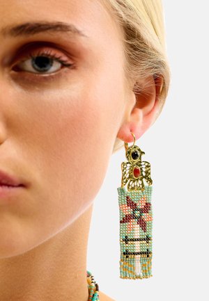 Boucles d'oreilles ton or avec un design d'oiseau comportant un œil et un accent rouge. Une frange perlée aux couleurs pastel avec des motifs géométriques pend en dessous.