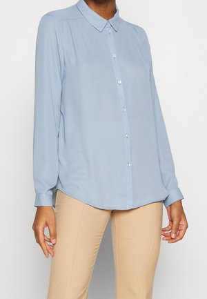 Personne portant une chemise bleu pâle à manches longues boutonnée et un pantalon beige, debout devant un fond uni.