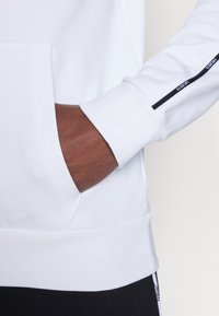 Hanorac alb cu un buzunar frontal, având o dungă neagră pe mânecă care afișează "Lacoste". Textură netedă cu manșete striate.