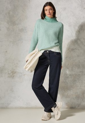 Muntgroene, geribbelde gebreide coltrui over een witte top, gecombineerd met donkerblauwe wijde jeans en beige sneakers. Zachte fleece-accessoire.