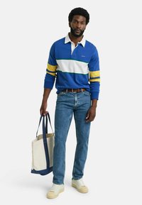 Blå langermet polo-shirt med hvite og gule striper, kombinert med blå jeans og kremfargede sneakers. En beige tote med marineblå detaljer holdes.