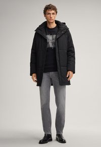 Giacca puffer nera con cappuccio, caratterizzata da un design trapuntato, chiusura con zip e indossata con una felpa nera casual sotto, abbinata a jeans grigi.