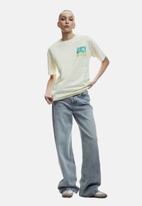 Kortærmet, bleg gul t-shirt med teksten "JUICY LEMON" i teal og olivengrøn, parret med højtaljede, lysblå, vide jeans.