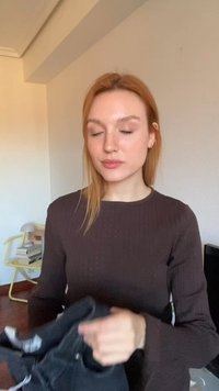 Jeune femme aux yeux fermés portant un pull foncé, tenant des vêtements noirs pliés dans une pièce avec une chaise jaune et des livres empilés.