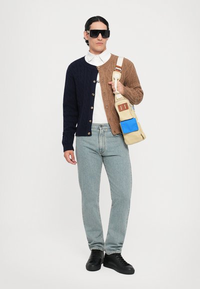Stickad cardigan med mörkblå och bruna sektioner, knappad fram, vit skjorta, ljusblå jeans och svarta sneakers. Bär en crossbody-väska.