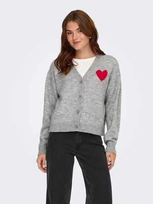 Femme portant un cardigan gris boutonné avec un cœur rouge sur la poitrine, une chemise blanche en dessous et un pantalon noir, debout devant un fond uni.