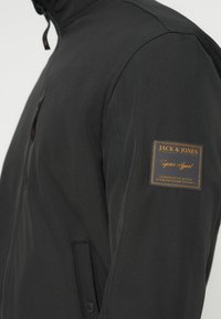 Μαύρο μπουφάν από υλικό με απαλή υφή, με φερμουάρ, μια τσέπη και πορτοκαλί λογότυπο στο μανίκι.