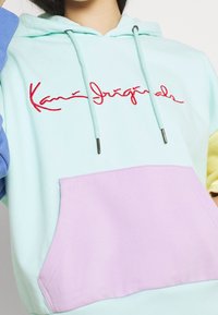 Mujer vestida con una sudadera de colores pastel en bloques, con un logo cursivo rojo en el pecho y un bolsillo frontal lavanda, con cordones azul claro visibles.