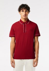 Röd pikétröja med korta ärmar, med krage och knappara, i kontrasterande vit sömnad. Emblem på vänster bröst.