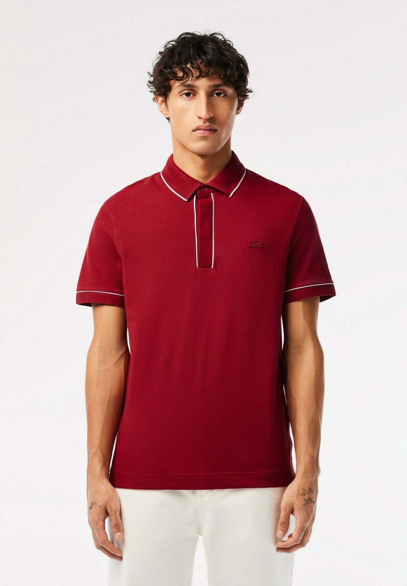 Röd pikétröja med korta ärmar, med krage och knappara, i kontrasterande vit sömnad. Emblem på vänster bröst.