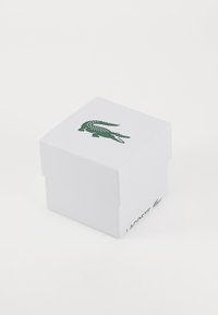 Baltas kartonas dėžutė su struktūrizuota paviršiumi, ant viršaus esantis žalias krokodilo logotipas ir šone spausdintas žodis "LACOSTE".