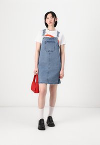 Denim kopējais kleita ar saplēstu apakšmalu, valkāta virs baltā t-kreklu. Aksesuāri ietver sarkanu rokassomu un melnu slip-on apavu pārī ar baltām zeķēm.