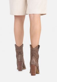Botas de tobillo trenzadas de color marrón con un acabado texturizado, tacón redondeado y un ligero corte abierto en los laterales. Diseño de tacón robusto y cónico.