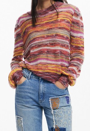 Person iført flerfarvet stribet strikket sweater med lange ærmer, stoppet ned i blå denimjeans med broderede lap-designs.