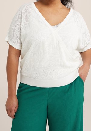 Kobieta ubrana w białą, krótkim rękawem, teksturowaną kopertową bluzkę na koronkowym topie na ramiączkach oraz zielone, teksturowane spodnie o szerokich nogawkach, ręka w kieszeni.