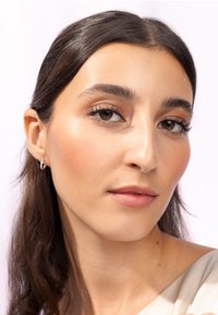 Acercamiento de un rostro con piel de oliva, cabello largo y oscuro, maquillaje natural y pendientes de aro dorados. La luz suave enfatiza la textura y los rasgos suaves.