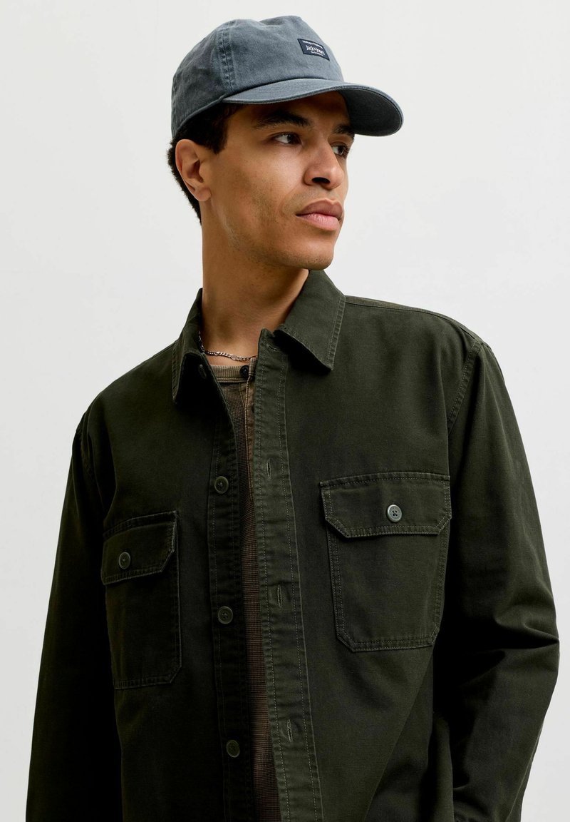 Jeune homme portant une chemise verte foncée à boutons et une casquette de baseball bleue, regardant vers sa gauche sur un fond clair uni.