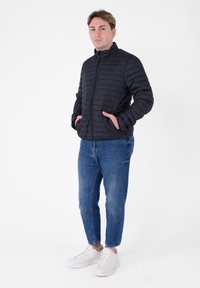 Zwarte pufferjas met een hoge kraag, ritssluiting en gewatteerde textuur; gecombineerd met blauwe jeans en witte sneakers.