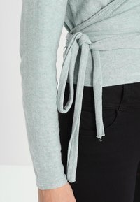 Jasnozielona bluzka z długim rękawem o kroju z oplataniem, związywana w talii. Materiał jest miękki i fakturowany, zestawiona z czarnymi spodniami.