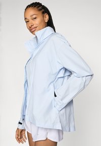 Chaqueta azul claro, resistente al agua, con un alto cuello, mangas largas y detalles negros en los puños y los lados. Cuenta con cierre de botones.
