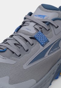 Grijze hardloopschoen met blauwe accenten, bovenwerk van mesh, platte veters, versterkte oogjes en een getextureerde rubberen zool, met "Timp 5" branding.