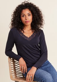Marine-blaues Langarmoberteil mit V-Ausschnitt und Spitzenbesatz. Weicher Stoff, lässige Passform, gezeigt mit blauen Jeans vor einem neutralen Hintergrund.