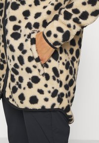 Krémová měkká bunda s dlouhým rukávem a černými leopardími skvrnami, která má boční kapsu a kontrastní černý lem podél okrajů.
