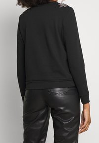 Svart sweatshirt med långa ärmar och ribbade muddar, med dekorativa detaljer på axlarna. Matchas med svarta byxor i slät textur.