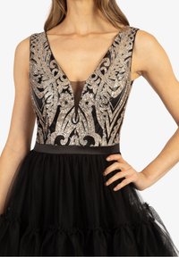 Robe noire avec un décolleté en V profond, ornée de perles en argent sur le corsage, et une jupe en tulle superposée avec un ourlet festonné.