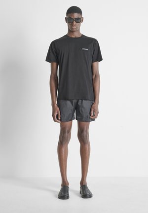 Jeune homme portant des lunettes de soleil noires, un t-shirt noir à manches courtes, un short noir et des chaussures noires sans lacets, debout devant un fond blanc uni.