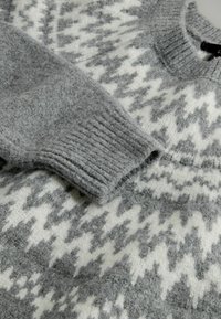 Grau-weißes Wollpullover mit Zickzackmuster, geripptem Bündchen und Kragen, flach ausgelegt mit dem Ärmel über die Brust gefaltet.