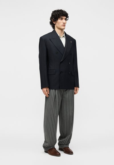 Άνδρας στέκεται φορώντας μαύρο διπλόστρωτο σακάκι, λευκό πουκάμισο, γκρι και λευκό ριγέ παντελόνι με φαρδιά πόδια και καφέ παπούτσια σε λευκό φόντο.