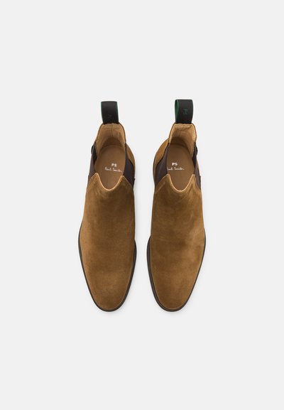 Bottines en suède marron avec des panneaux élastiques sur les côtés et des languettes de tirage. Présente une texture lisse et une pointe arrondie. Design minimaliste.