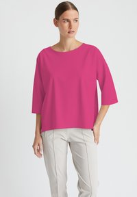 Fuchsia oversized Oberteil mit Rundhalsausschnitt und Dreiviertelärmeln, hergestellt aus glattem Stoff, kombiniert mit hellen Hosen.