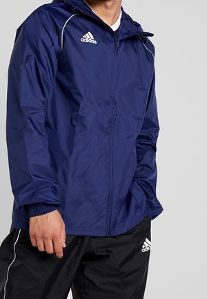 Mann trägt eine marineblaue Adidas Reißverschlussjacke und schwarze Adidas Trainingshose, die Hände in den Taschen, vor einfachem Hintergrund.