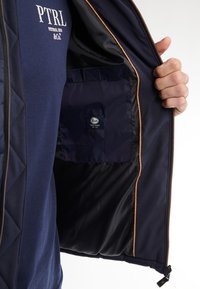 Chaqueta navy con exterior acolchado, forro naranja contrastante y bolsillo interior con etiqueta de marca. Cierre con cremallera en el frente.
