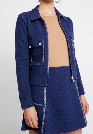 Femme portant une veste bleue texturée avec des poches poitrine à boutons et une jupe assortie, associée à un haut beige et de grandes boucles d'oreilles.
