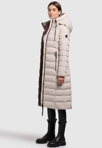 Dlhý, svetlo béžový pufový kabát s kapucňou, s vertikálnym prešívaním, ktorý má čierne kovové doplnky a marhuľovú podšívku.