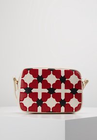 Crossbody bag features a cream base with bold red heart patterns, black accents, and gold-tone hardware. Smooth texture with a structured shape.