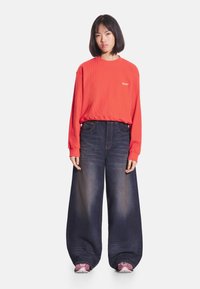 Czerwona prążkowana bluza z długim rękawem z logo, zestawiona z luźnymi, ciemnoniebieskimi dżinsami. Modelka ma na nogach różowe trampki.