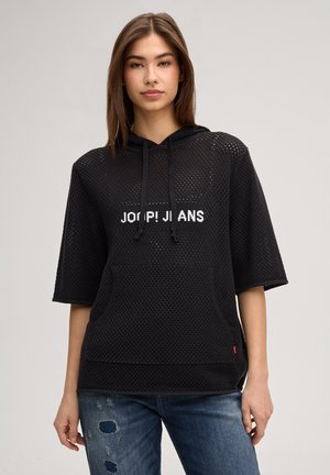 Giovane donna che indossa una felpa nera a rete con la scritta "JOOP! JEANS" e jeans blu strappati, in piedi davanti a uno sfondo semplice.