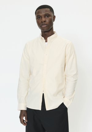 Hellgelbes gestreiftes Hemd mit Stehkragen, durchgehendem Knopfleiste und langen Ärmeln. Der Stoff ist glatt; kombiniert mit dunkler Hose für Kontrast.