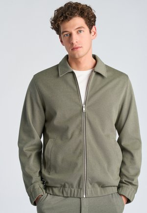 Jonge man met krullend haar draagt een licht olijfgroen jasje met rits en bijpassende broek, staand tegen een effen lichte achtergrond.