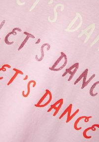 Camiseta de algodón de color rosa claro con el texto "¡BAILEMOS!" en tamaños y colores variados: magenta brillante y rojo intenso. Textura suave.