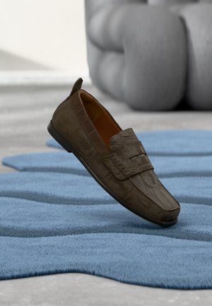 Mocassin en cuir marron avec motif embossé posé sur un tapis bleu texturé, mobilier gris clair flou en arrière-plan.