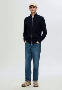 Suéter de cremallera azul marino, jeans de mezclilla azul claro, sandalias deslizables beige y una gorra beige llevada por una figura masculina en un fondo neutro.