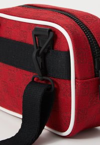 Rote strukturierte Stofftasche mit weißen Akzenten und einem schwarzen verstellbaren Tragegurt. Ausgestattet mit einem schwarzen Reißverschluss und einem praktischen Clip-Verschluss.