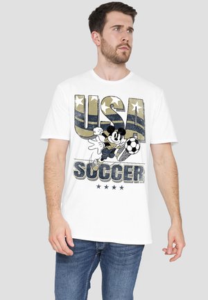 Mann trägt weißes T-Shirt mit dem Schriftzug "USA Soccer" und einer Cartoon-Maus, die einen Fußball in Rot, Weiß und Blau kickt.