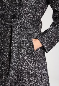 Svart och vit prickig kappa med bälte i midjan, stora knappar och sidfickor; tillverkad av strukturerad tyg.